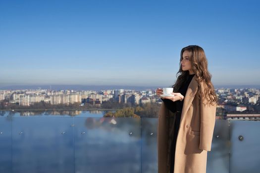 Side view of adorable young woman standing on balcony with cup of coffee and enjoying amazing view on big city. Concept of morning time and lifestyle.