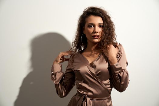 Portrait of attractive woman with curly brown hair posing near white wall, touching shoulders. Front view of beautiful seductive young female model wearing beige silk dress. Concept of beauty.