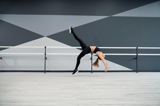 Side view of strong fit woman practicing wheel in jump, doing split in air. Attractive flexible sportswoman training in dance hall with handrails. Concept of choreography, healthy lifestyle.