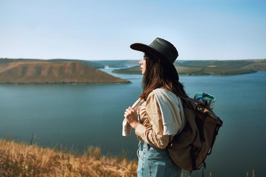 Positive young woman in cowboy hat walking among green nature at Bakota area. Happy female with backpack spending free time actively.