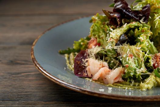 Close up view of tasty light salad with lettuce and shrimps covered with balsamic dressing. Healthy snack with seafood and cheese on blue plate on wooden table in luxury restaurant.