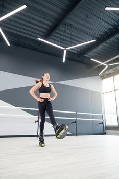 Young caucasian girl with ponytail practicing kangoo jumping indoors. From below view of female athlete holding hands on waist, enjoying cardio dance workout in hall, hi tech interior.