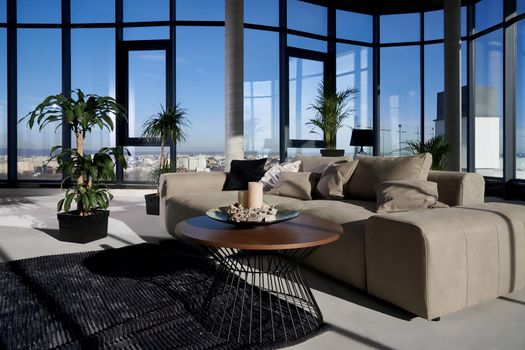 Front view of fashionable interior spacious room with large panoramic window with incredible view on city and cozy grey sofa with pillows and green flowerpots. Concept of modern interior.