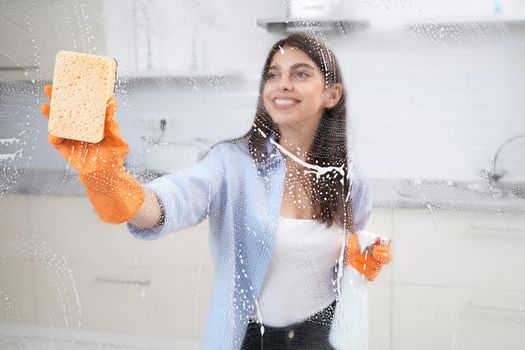 Smiling brunette woman washing window in rubber gloves with rag and detergent. Concept of cleaning window or glass.