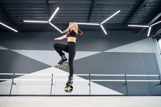 From below view of active female teenager doing cardio workout in hall, hi tech interior. Young pretty girl wearing black sportswear practicing dance moves while doing kangoo jumps and smiling.