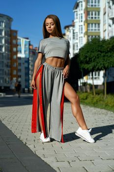Front full length view of young fashionable woman posing outdoors in urban summer atmosphere. Female model wearing extraordinary sports trousers with naked legs standing near multistorey buildings.