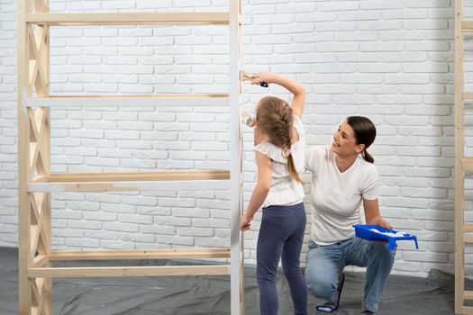 Smiling young woman with daughter painting wooden rack in white color at home. Concept of process painting.