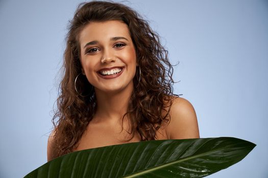 Portrait of young woman with perfect makeup laughing, hiding chest behind big green leaf. Attractive sincere naked female model with curly hair and big earrings looking at camera isolated on blue.