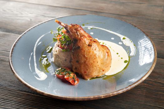 Side view of juicy roasted bbq chicken leg served with mashed potato and mushrooms, cherry tomatoes, decorated with fresh green sprouts, sour cream and green sauce. Blue ceramic plate on wooden table.