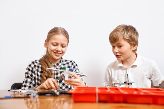 Interesting building kit for kids on table. Front view of boy and girl having fun, creating air vehicle. Science engineering. Nice interested friends smiling, chatting and working on project together.