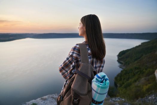 Adorable dark-haired woman in checkered shirt enjoying amazing view of summer sunset at Bakota bay. Young hiker with backpack standing on mountain peak.