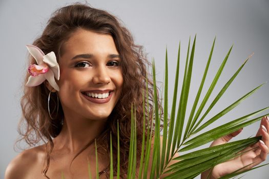 Pretty happy female model with naked shoulders and curly brown hair looking at camera and holding palm leaf. Young woman with perfect makeup laughing, posing with pink orchid flower behind ear.