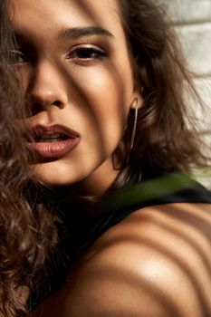 Crop of seductive young brunette female model with mouth open looking at camera. Close up portrait of pretty woman with curly hair posing near white brick wall, shadows on face. Concept of beauty.