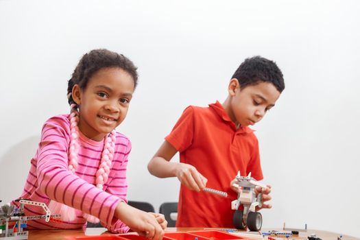 Front view of building kit for group of kids creating toys, having positive emotions and joy, selective focus of girl smiling and looking at camera. Close up of african friends working on project.