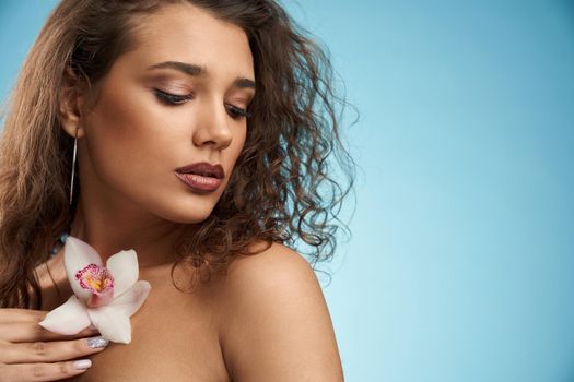 Crop of young attractive brunette female model holding pink and white orchid on naked chest and looking aside. Sensual woman with curly hair posing holding flower in hand. Beauty, modeling concept.