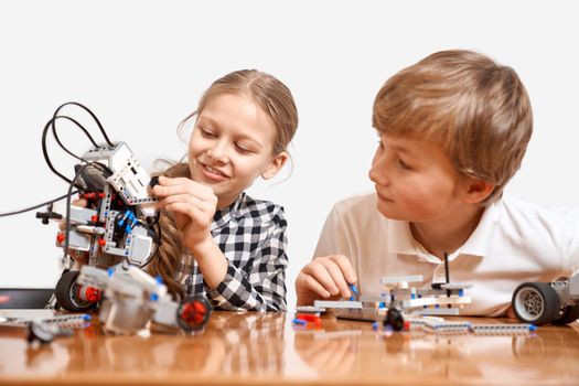 Front view of kids having fun, creating toys. Science engineering. Nice interested friends smiling, chatting, girl showing boy robot made with interesting building kit for kids on table.