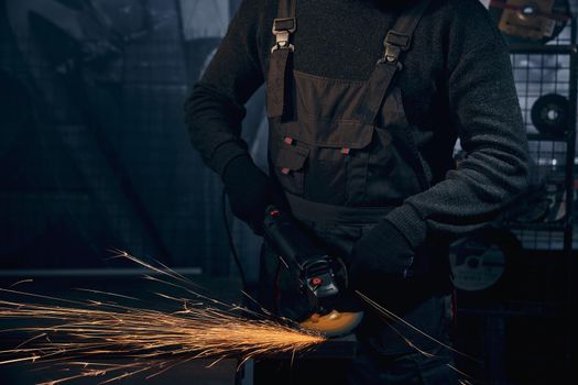 Close up of worker in special black suit and gloves working with special equipment for metal. Concept of man polishing metal in dark room or garage with angle grinder.