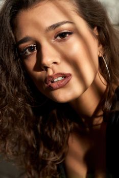 Beautiful seductive young female model wearing black dress. Close up portrait of gorgeous woman with curly brown hair posing near white brick wall, looking at camera, shadows. Concept of beauty.