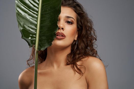 Portrait of young naked woman with perfect makeup posing with mouth open and hiding half of face behind green leaf. Close up of beautiful female model standing and looking at camera isolated on gray.