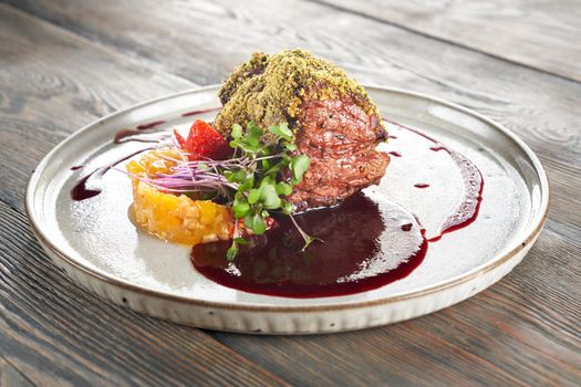 Close up view of beef meat with pistachio topping served with cranberry sauce. Tasty meal with fruits, berries and fresh sprouts presented on ceramic white plate in luxurious restaurant, wooden table.
