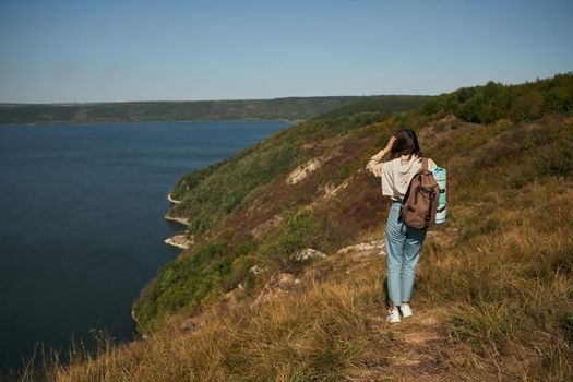 Back view of young woman with backpack walking along high green hill at Dniester canyon. Female traveler discovering beautiful places in Ukraine during hiking alone.