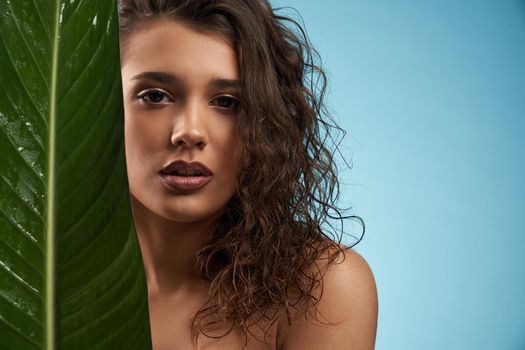 Close up of beautiful naked female model looking at camera. Crop portrait of young brunette woman with perfect makeup posing with mouth open, holding big green leaf, isolated on blue.