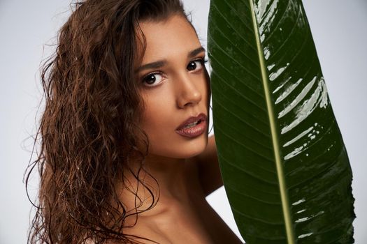 Portrait of young woman with perfect makeup posing with mouth open behind big green leaf. Side view of attractive brunette naked female model posing and looking at camera isolated on white.