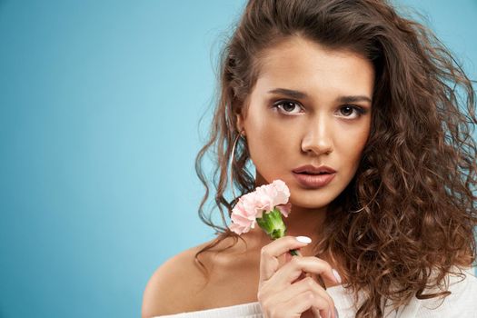 Close up of fashionable brunette model with naked shoulders looking at camera. Pretty young woman with perfect makeup and curly hair posing with pink clove flower in hand isolated on blue background.