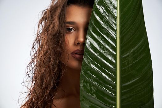 Close up of beautiful female model standing and looking at camera isolated on white. Portrait of young naked woman with perfect makeup posing with mouth open and hiding half of face behind green leaf.