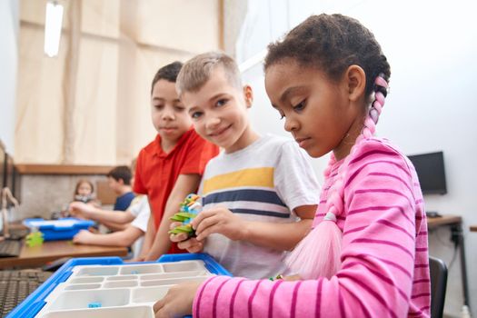 Side view of building kit with colorful peices in box for group of three multiracial kids creating toys. Close up of interested friends working on project, having positive emotions and joy.