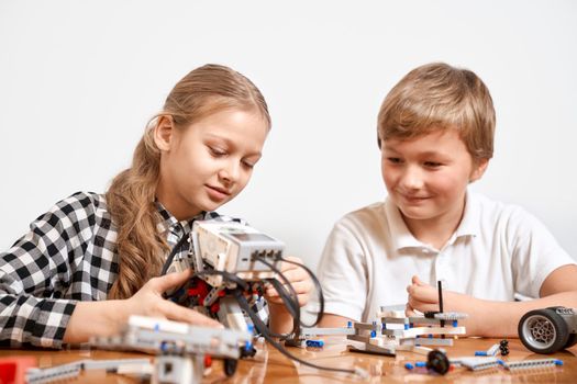 Front view of boy and girl having fun, creating robot. Science engineering. Nice interested friends smiling, chatting and working on project together using interesting building kit for kids on table.