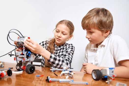 Front view of boy helping girl in creating robot using building kit for kids on table. Nice interested friends smiling, chatting and working on project together. Concept of science engineering.