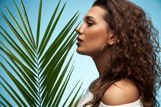 Side view of sensual brunette female model with naked shoulders profile headshot. Pretty young woman with perfect fancy makeup posing near green big palm leaf isolated on blue studio background.