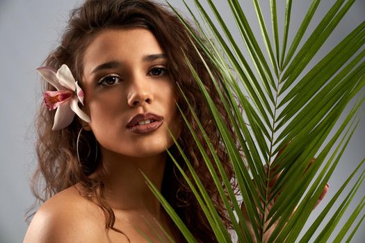 Female model with naked shoulders and curly brown hair looking at camera, holding palm leaf. Young gorgeous woman with perfect makeup posing with orchid flower behind ear isolated on gray background.