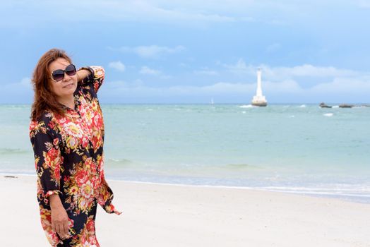 Middle-aged woman tourist wearing sunglasse looking at the camera and smiling on the beach and sea in summer sky background at Koh Tarutao island, Satun, Thailand