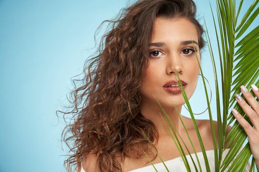 Portrait of attractive brunette female model wearing naked shoulders dress looking at camera with mouth open. Pretty young woman with perfect makeup posing with green big palm leaf isolated on blue.
