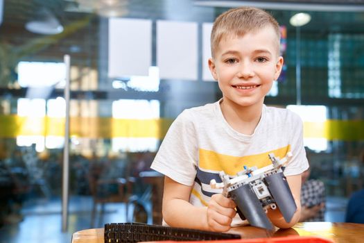 Front view of lovely caucasian boy smiling and looking at camera. Building kit for kids on table, children creating toys, having positive emotions and joy. Close up of boy working on project.