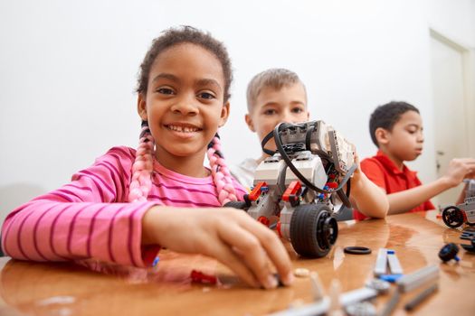 Front view of building kit for group of three multiracial kids creating toys, having positive emotions and joy, african girl smiling and looking at camera. Close up of friends working on project.