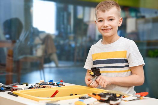 Front view of lovely caucasian boy smiling and looking at camera. Building kit for kids on table, children creating toys, having positive emotions and joy. Close up of boy working on project.