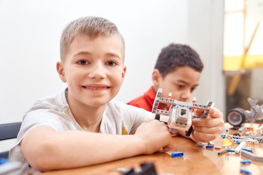 Side view of building kit for group of multiracial kids creating toys, having positive emotions and joy, lovely caucasian boy smiling and looking at camera. Close up of friends working on project.