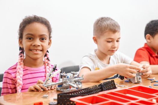 Side view of building kit for group of three multiracial kids creating toys, having positive emotions and joy, african girl smiling and looking at camera. Close up of friends working on project.
