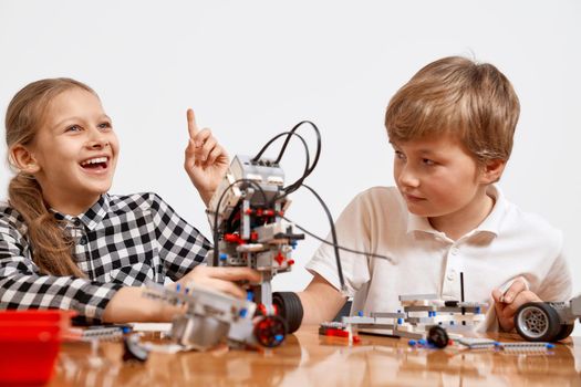 Front view of boy and girl having fun, creating vehicle. Science engineering. Friends working on project together, girl having idea and pointing with finger up. Building kit for kids on table.