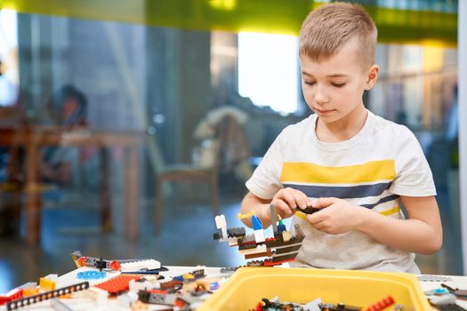 Selective focus of lovely caucasian boy working on project, taking colorful parts to create robot, having positive emotions and joy. Front view of building kit for kids. Science engineering.