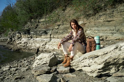 Pretty young woman dressed in casual clothing sitting on stone near Dniester river. Female hiker with backpack relaxing during active time spending on fresh air.
