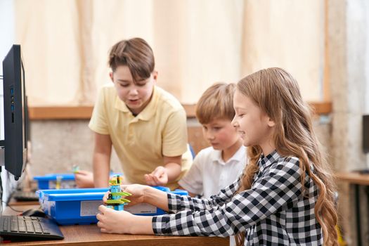 Interesting building kit for kids on table with computers. Side view of boys and girl creating toys. Science engineering. Nice interested friends chatting and working on project together.