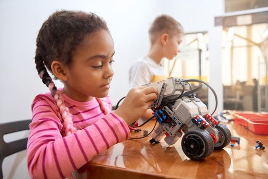 Building kit with colorful pieces for group of multiracial kids creating toys. Close up of concentrated african girl working on project, having positive emotions.. Concept of science engineering.