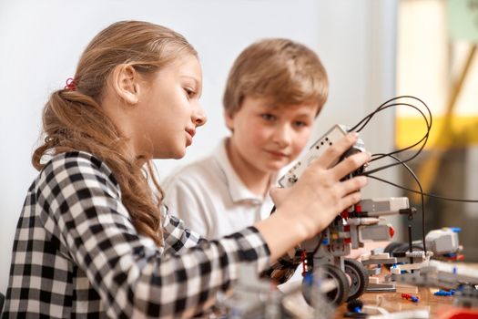 Side view of boy helping girl in creating robot using building kit for kids on table. Nice interested friends smiling, chatting and working on project together. Concept of science engineering.