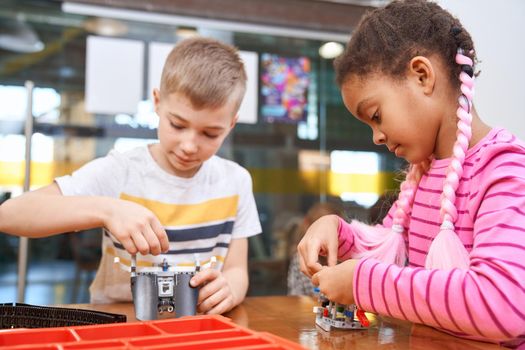 Building kit for group of multiracial kids creating toys. Close up of concentrated african girl and caucasian boy working on project, having positive emotions. Concept of science engineering.