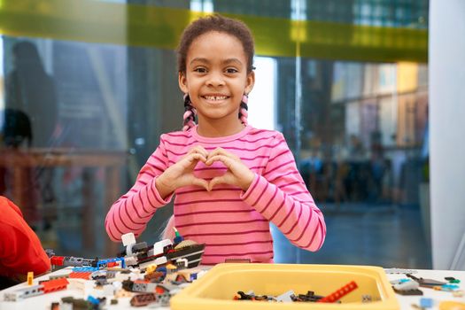 Front view of lovely smiling african girl standing, looking at camera and making heart with hands. Building kit for kids on table, children creating toys, having positive emotions and joy.