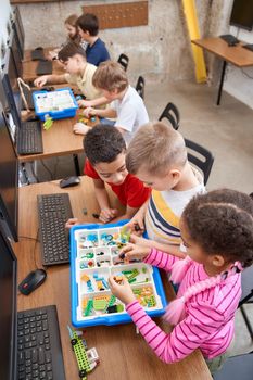 Interesting building kit for kids on table with computers. From above view of boys and girls creating toys. Science engineering. Nice interested friends chatting and working on project together.